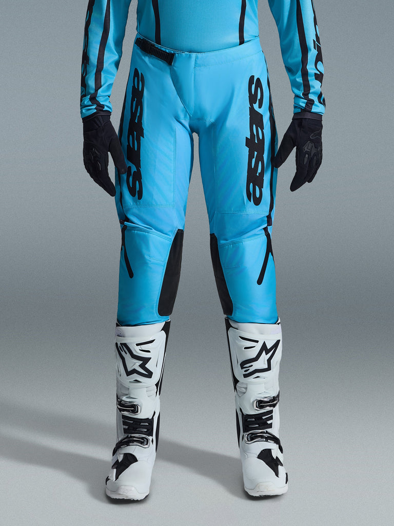 Alpinestars Stella Fluid Apex 2026, damskie spodnie motocrossowe, jasnoniebieskie i czarne, pionowe czarne logo na udach, czarne wzmocnione panele po wewnętrznej stronie kolan, widoczne z biało-czarnymi butami motocrossowymi i czarnymi rękawicami, profesjonalna odzież off-road dla kobiet