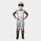 Zawodniczka motocrossowa w spodniach Alpinestars Stella Techstar 2024, kolor Black Multicolor z białą bazą i czerwonymi, żółtymi oraz niebieskimi pasami, dopasowana koszulka z długim rękawem, biały kask i czarne buty, pełny strój wyścigowy do zawodów terenowych