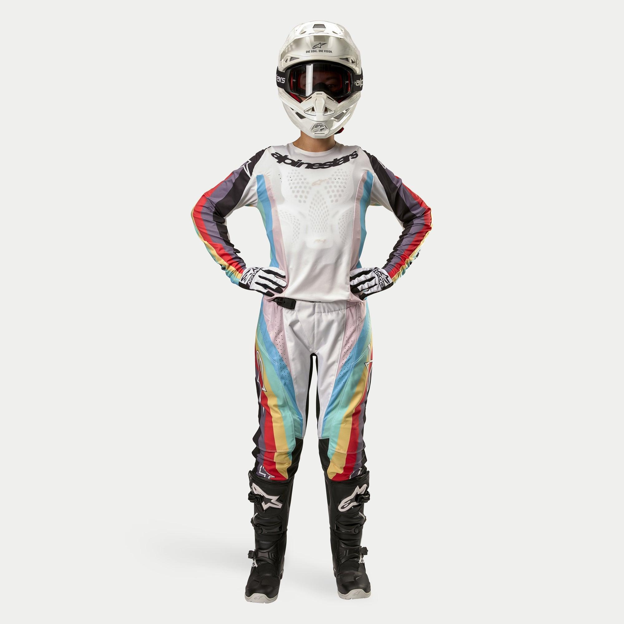 Zawodniczka motocrossowa w spodniach Alpinestars Stella Techstar 2024, kolor Black Multicolor z białą bazą i czerwonymi, żółtymi oraz niebieskimi pasami, dopasowana koszulka z długim rękawem, biały kask i czarne buty, pełny strój wyścigowy do zawodów terenowych