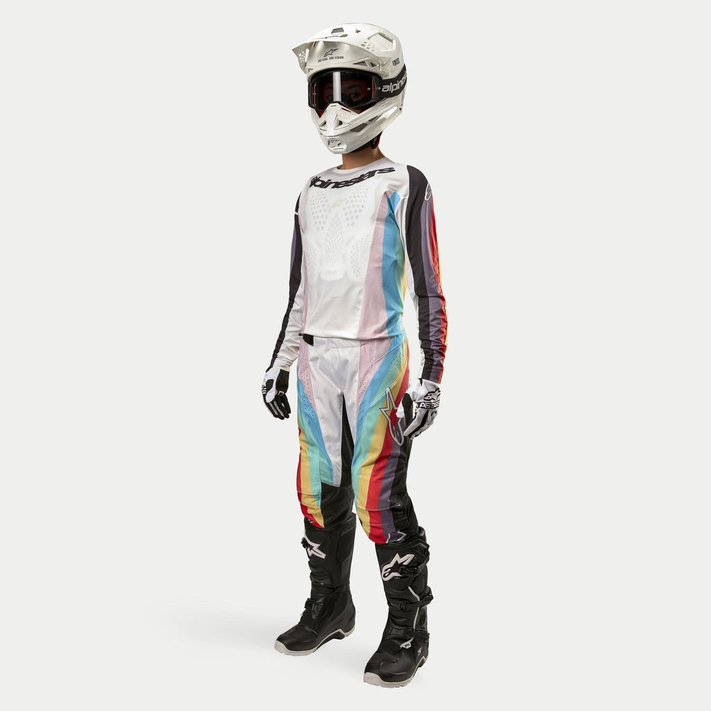 Zawodniczka motocrossowa w damskich spodniach Alpinestars Stella Techstar 2024, spodnie motocrossowe, kolorystyka Black Multicolor z białymi panelami i wyrazistymi ukośnymi pasami, pasująca koszulka, biały kask i czarne buty, kompletny damski strój do wyścigów terenowych