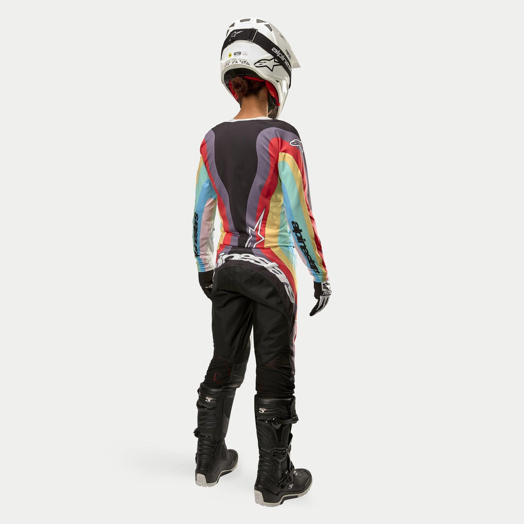 Motocyklistka motocrossowa widoczna od tyłu w damskich spodniach Alpinestars Stella Techstar 2024 Black Multicolor, duże białe logo na siedzeniu, kolorowa koszulka w paski, biały kask i czarne buty Tech 3 do wyścigów off-road