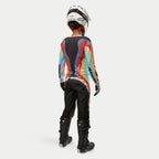 Motocyklistka motocrossowa widoczna od tyłu w damskich spodniach Alpinestars Stella Techstar 2024 Black Multicolor, duże białe logo na siedzeniu, kolorowa koszulka w paski, biały kask i czarne buty Tech 3 do wyścigów off-road