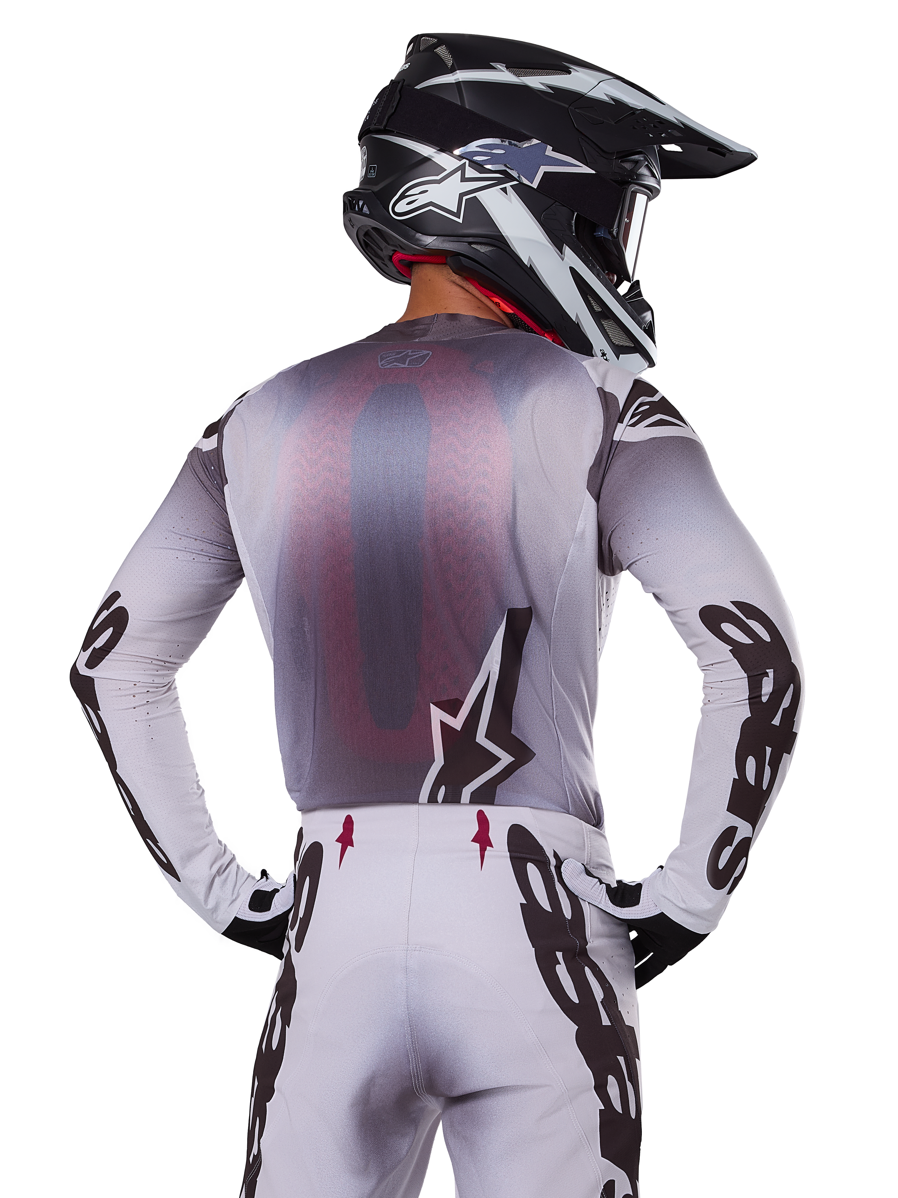 Zestaw odzieży Alpinestars 2025 Supertech Maker, motocyklista motocrossowy widoczny od tyłu, biało-szara koszulka z dużym czarnym brandingiem na rękawach i logo gwiazdy, pasujące białe spodnie z czarną grafiką, czarno-biały kask, profesjonalny strój do wyścigów terenowych