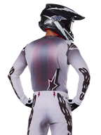 Motocyklista motocrossowy widziany od tyłu w koszulce Alpinestars 2025 Supertech Maker, srebrno-czarna z czerwonym siatkowym panelem na plecach i czarnymi logotypami na rękawach, w zestawie z dopasowanymi spodniami motocrossowymi i czarno-białym kaskiem, profesjonalny sprzęt wyścigowy off-road