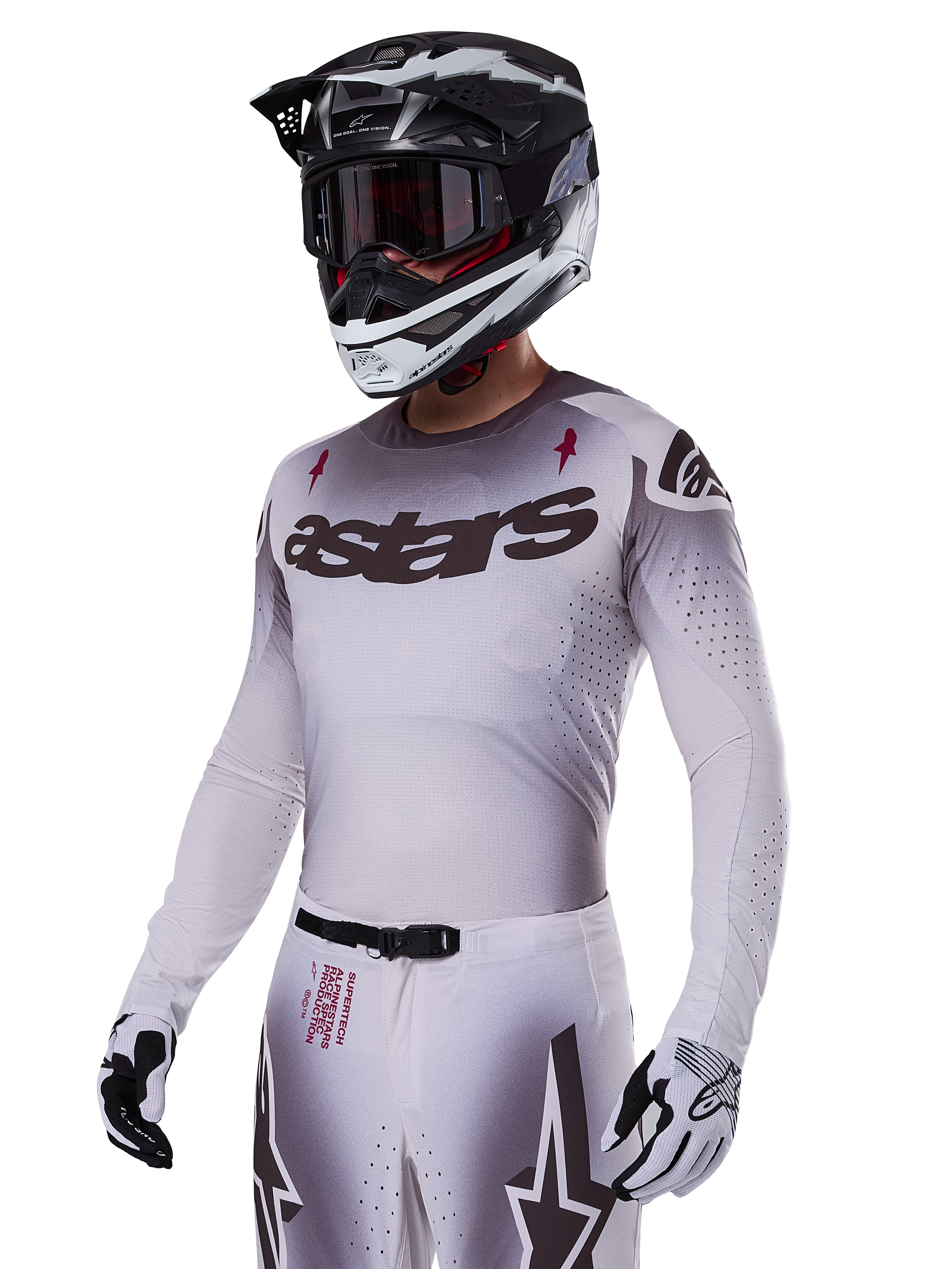 Motocyklista motocrossowy w zestawie Alpinestars 2025 Supertech Maker w kolorze białym, szara koszulka z gradientem, czarnym logo astars i laserowymi perforacjami, pasujące spodnie z klamrą magnetyczną i grafiką gwiazdy, czarno-biały kask i rękawice, kompletny strój wyścigowy off-road