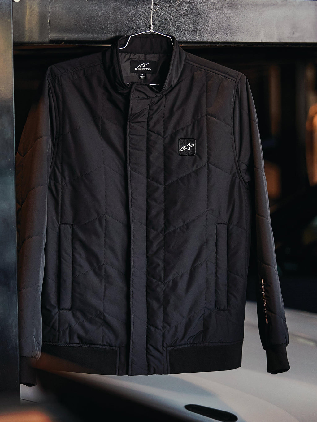 Alpinestars Precedent Jacket, pikowana kurtka sportowa, czarna, ze skośnym wzorem pikowania, niskim kołnierzem torowym z zakrytym zamkiem, naszywką z logo na piersi oraz ściągaczami przy mankietach i na dole, design inspirowany sportami motorowymi