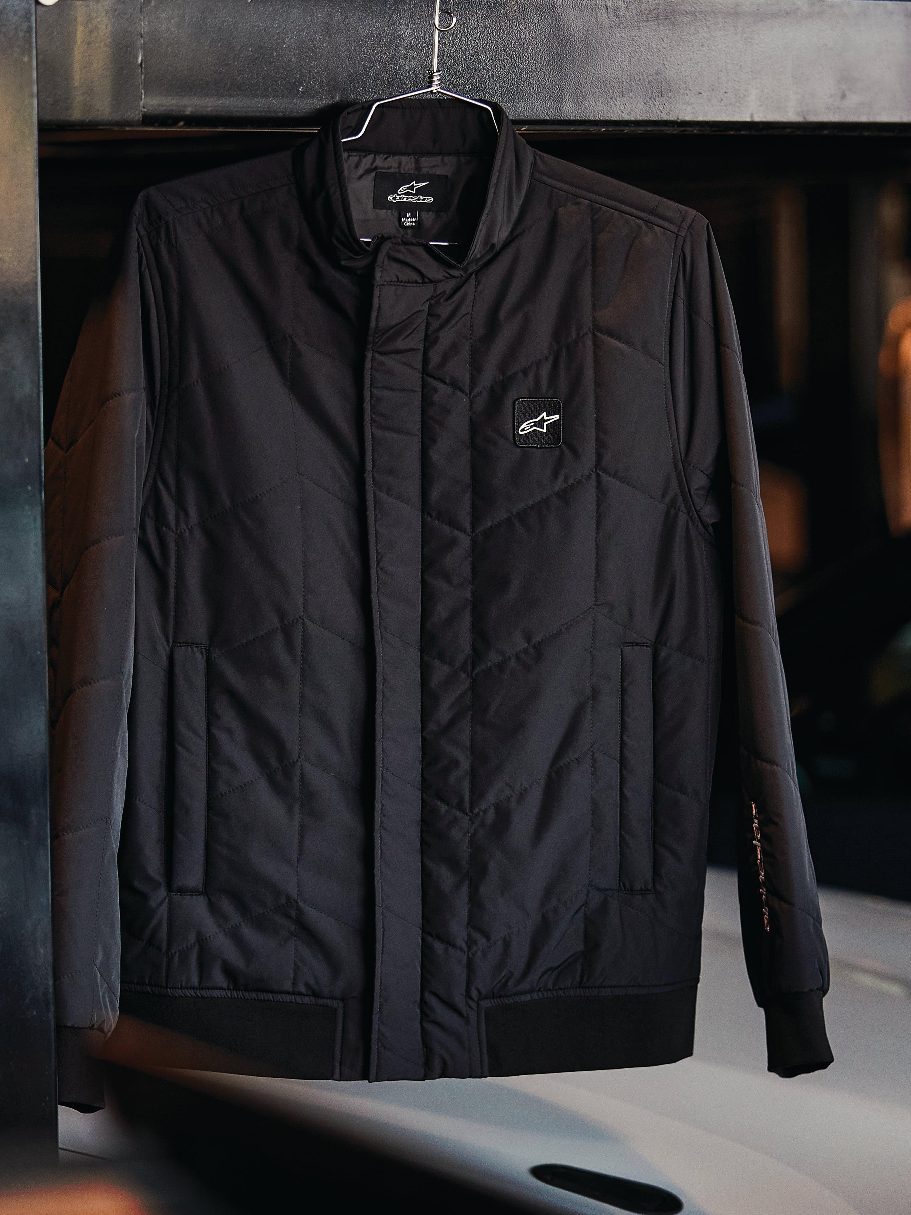 Alpinestars Precedent Jacket, pikowana kurtka sportowa, czarna, ze skośnym wzorem pikowania, niskim kołnierzem torowym z zakrytym zamkiem, naszywką z logo na piersi oraz ściągaczami przy mankietach i na dole, design inspirowany sportami motorowymi