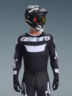 Alpinestars 2026 Racer Air Riway Jersey, czarno-biała koszulka motocrossowa z brandingiem „astars”, noszona przez motocyklistę w pasującym czarnym kasku, goglach i spodniach, z oddychającymi panelami z siatki do wyścigów terenowych.