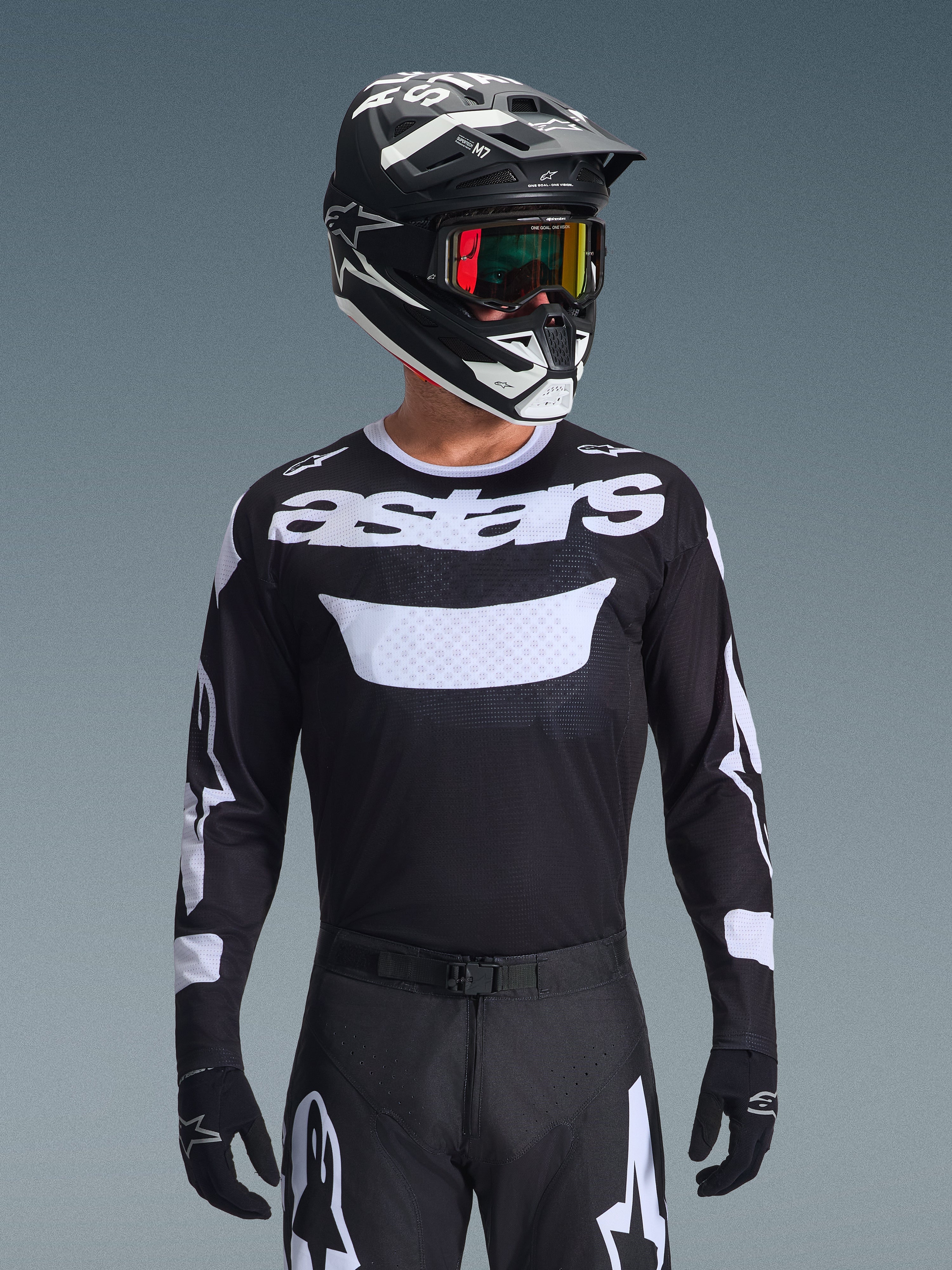 Zawodnik motocrossowy w zestawie Alpinestars 2026 Racer Air Riway, czarna koszulka z białym logo „astars” i wentylowanym panelem na klatce piersiowej, czarne spodnie z białymi akcentami, dopasowany czarno-biały kask z opalizującymi goglami, czarne rękawice, wysokowydajny strój motocrossowy w czarno-białej kolorystyce.