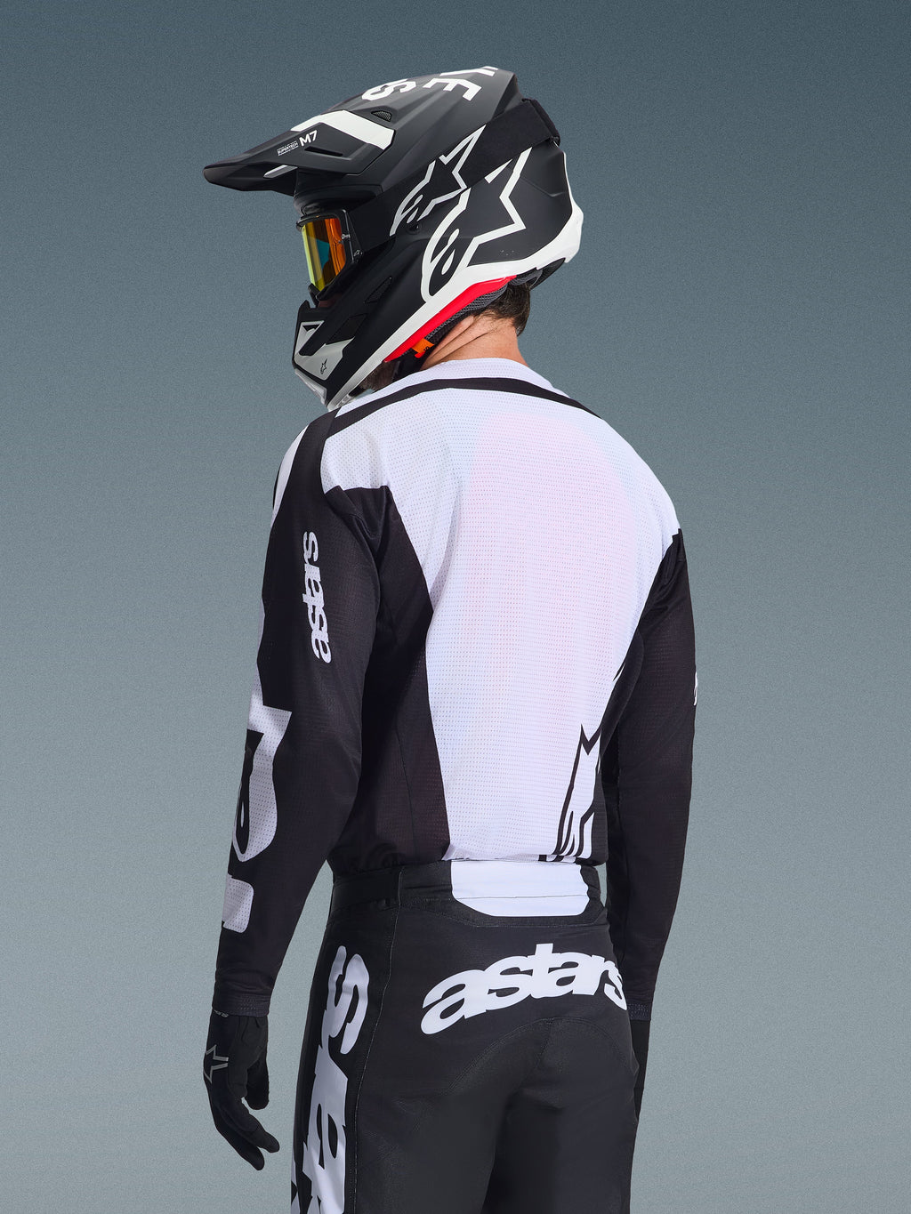 Motocyklista motocrossowy widziany od tyłu w koszulce Alpinestars 2026 Racer Air Riway, czarno-biała, z białym oddychającym panelem z siateczki na plecach i czarnymi rękawami z brandingiem „astars”, pasujące czarne spodnie z białym logo, czarny kask z białymi akcentami, profesjonalny sprzęt off-road o wysokiej wydajności w wysokich temperaturach.