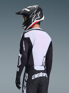 Motocyklista motocrossowy widziany od tyłu w koszulce Alpinestars 2026 Racer Air Riway, czarno-biała, z białym oddychającym panelem z siateczki na plecach i czarnymi rękawami z brandingiem „astars”, pasujące czarne spodnie z białym logo, czarny kask z białymi akcentami, profesjonalny sprzęt off-road o wysokiej wydajności w wysokich temperaturach.