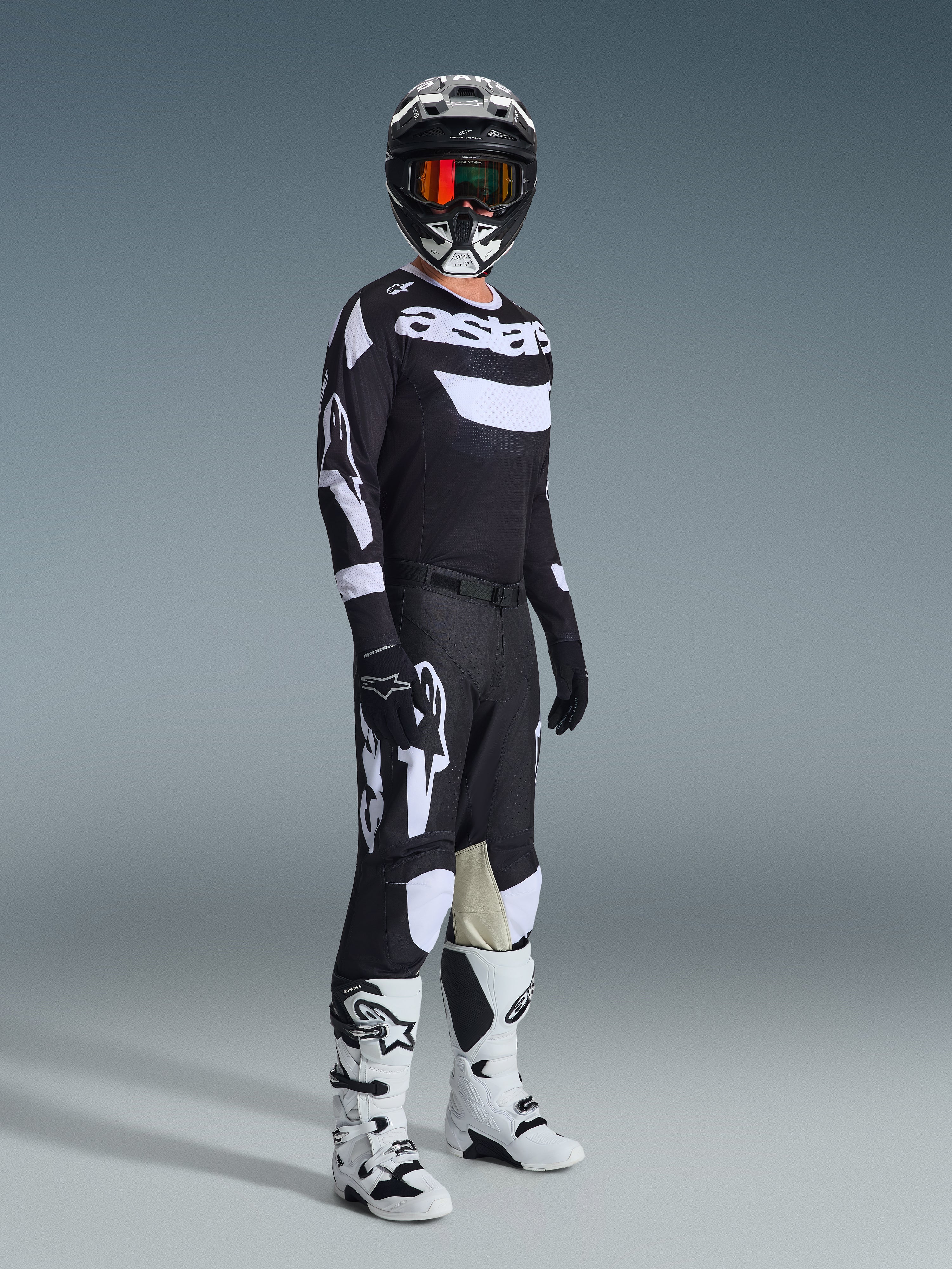 Zestaw Alpinestars 2026 Racer Air Riway, zawodnik motocrossowy w pełnym stroju wyścigowym, czarna koszulka z białym brandingiem „Astars” i panelami z siatki, czarne spodnie z dużymi białymi logo i panelami na kolanach, czarny kask z czerwonymi goglami, biało-czarne buty, profesjonalny strój do wyścigów off-road.
