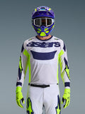 Alpinestars 2026 Racer Air Riway Jersey, jasnoszara koszulka motocrossowa z ciemnoniebieskim logo astars, neonowożółte i niebieskie akcenty na rękawach, noszona przez motocyklistę z dopasowanym fioletowym i neonowożółtym kaskiem oraz rękawicami, techniczna tkanina siatkowa do wyścigów off-road
