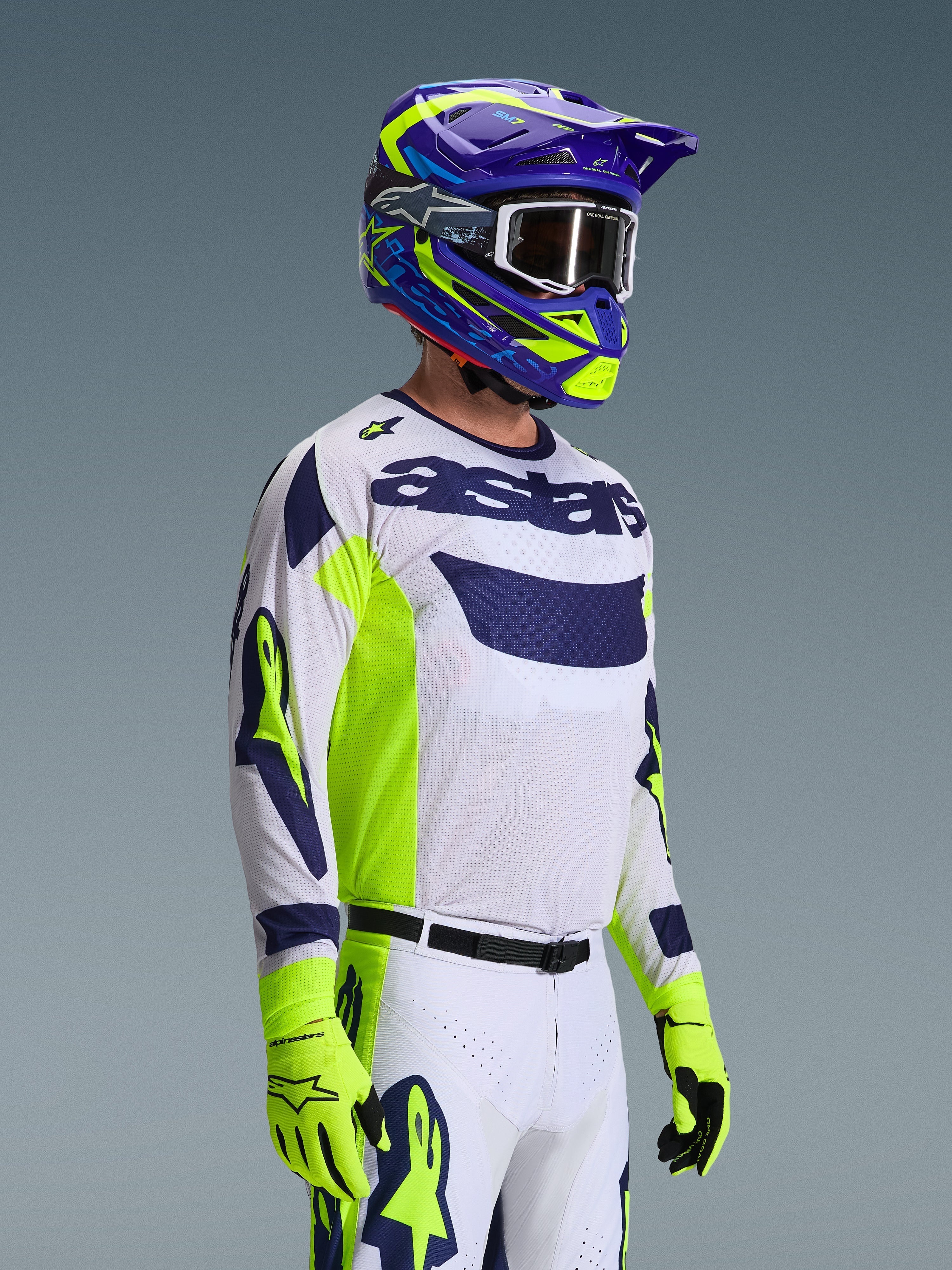 Motocyklista motocrossowy w koszulce Alpinestars 2026 Racer Air Riway, jasnoszara z akcentami w kolorze żółtym fluo i niebieskim, w zestawie z dopasowanym niebiesko-żółtym kaskiem i jaskrawożółtymi rękawicami, oddychająca konstrukcja z siateczki, profesjonalna stylistyka wyścigowa do jazdy off-road.