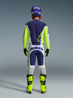 Widok z tyłu zawodnika motocrossowego w koszulce Alpinestars 2026 Racer Air Riway i pasujących spodniach w kolorze jasnoszarym, żółtym fluo i niebieskim, z jaskrawożółtymi butami fluo i dopasowanym kaskiem, prezentujący kompletny zestaw odzieży wyścigowej off-road.