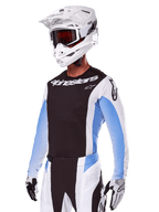 Motocyklista motocrossowy w zestawie Alpinestars 2025 Techstar Melt, biały z czarnymi i jasnoniebieskimi akcentami, koszulka z czarnym przodem i białymi rękawami, pasujące biało-czarne spodnie, biały kask SM10 z goglami i czarno-białe rękawice, kompletny profesjonalny strój wyścigowy off-road.