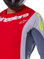 Koszulka Alpinestars Techstar Melt 2025, koszulka motocrossowa, jasnoczerwona i szara z ciemnoniebieskimi panelami, białe logo Alpinestars i neonowożółte logo na ramieniu, zbliżenie na laserowo wycinane otwory wentylacyjne, czarny kask