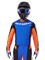 Koszulka motocrossowa Alpinestars Techstar Melt 2025, niebiesko-pomarańczowa z czarnymi panelami bocznymi, noszona z czarnym kaskiem i niebieskimi goglami, posiada laserowo wycinane perforacje na klatce piersiowej, sportowy krój wyścigowy do jazdy terenowej