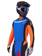 Zestaw Alpinestars 2025 Techstar Melt, zawodnik motocrossowy w niebieskiej koszulce z pomarańczowymi rękawami i czarnymi panelami bocznymi, dopasowane niebiesko-czarne spodnie, czarny kask z niebieskimi goglami, biało-czarne rękawice, sportowa odzież motocrossowa o kroju wyścigowym