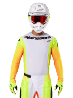 Alpinestars 2025 Techstar Melt Jersey, odzież do wyścigów motocrossowych, biały przód z neonowo-żółtymi i pomarańczowymi rękawami, zawodnik w białym kasku i czerwonych goglach, neonowo-żółte rękawice z czarnym logo, wyrazista kolorystyka dla wydajności off-road