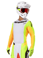 Motocyklista motocrossowy w zestawie Alpinestars 2025 Techstar Melt, biała koszulka z jaskrawymi żółtymi i pomarańczowymi rękawami, czarne logo Alpinestars na klatce piersiowej, dopasowane neonowożółte rękawice, biały kask z goglami z czerwonymi szkłami, profesjonalny zestaw do wyścigów terenowych