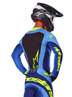 Widok z tyłu zawodnika motocrossowego ubranego w koszulkę Alpinestars 2025 Techstar Melt w kolorze niebieskim i neonowym żółtym, z czarnym panelem na plecach i wyraźnym żółtym logo na rękawach, w zestawie z dopasowanymi spodniami motocrossowymi i czarnym kaskiem karbonowym, profesjonalna odzież wyścigowa off-road