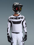 Zawodnik motocrossowy w koszulce Alpinestars 2026 Racer Riway w kolorze biało-czarnym, z logo „astars” na klatce piersiowej, w zestawie z pasującymi biało-czarnymi spodniami, czarnym kaskiem z goglami i czarnymi rękawicami, kompletny strój do jazdy terenowej