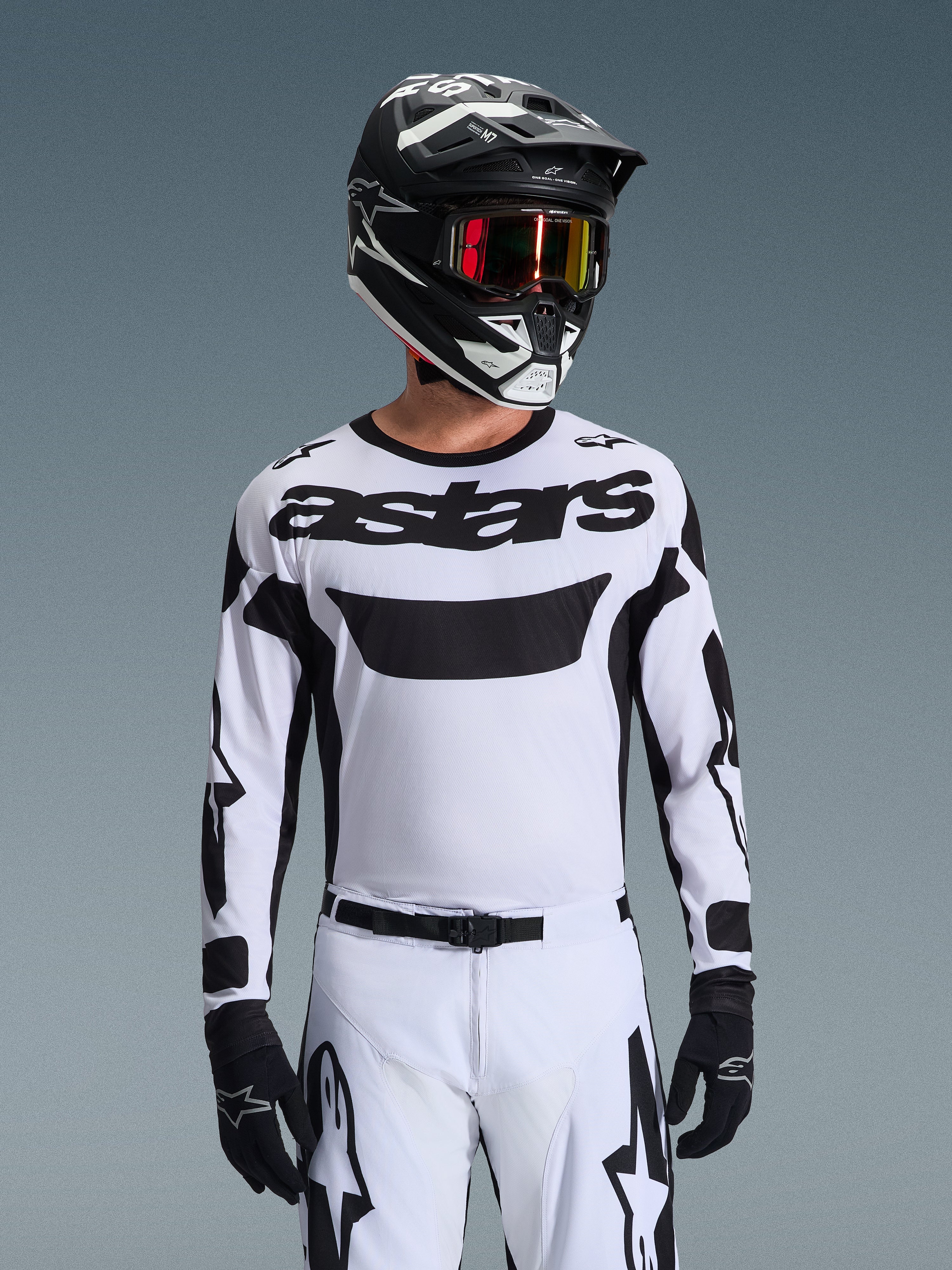 Zawodnik motocrossowy w koszulce Alpinestars 2026 Racer Riway w kolorze biało-czarnym, z logo „astars” na klatce piersiowej, w zestawie z pasującymi biało-czarnymi spodniami, czarnym kaskiem z goglami i czarnymi rękawicami, kompletny strój do jazdy terenowej