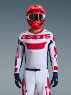 Zawodnik motocrossowy w koszulce Alpinestars 2026 Racer Riway w kolorystyce biało-niebiesko-czerwonej, duże czerwone logo „astars” na piersi, granatowe panele boczne, dopasowane biało-czerwone spodnie, czerwony kask z białymi goglami i granatowe rękawice, kompletny zestaw wyścigowy off-road