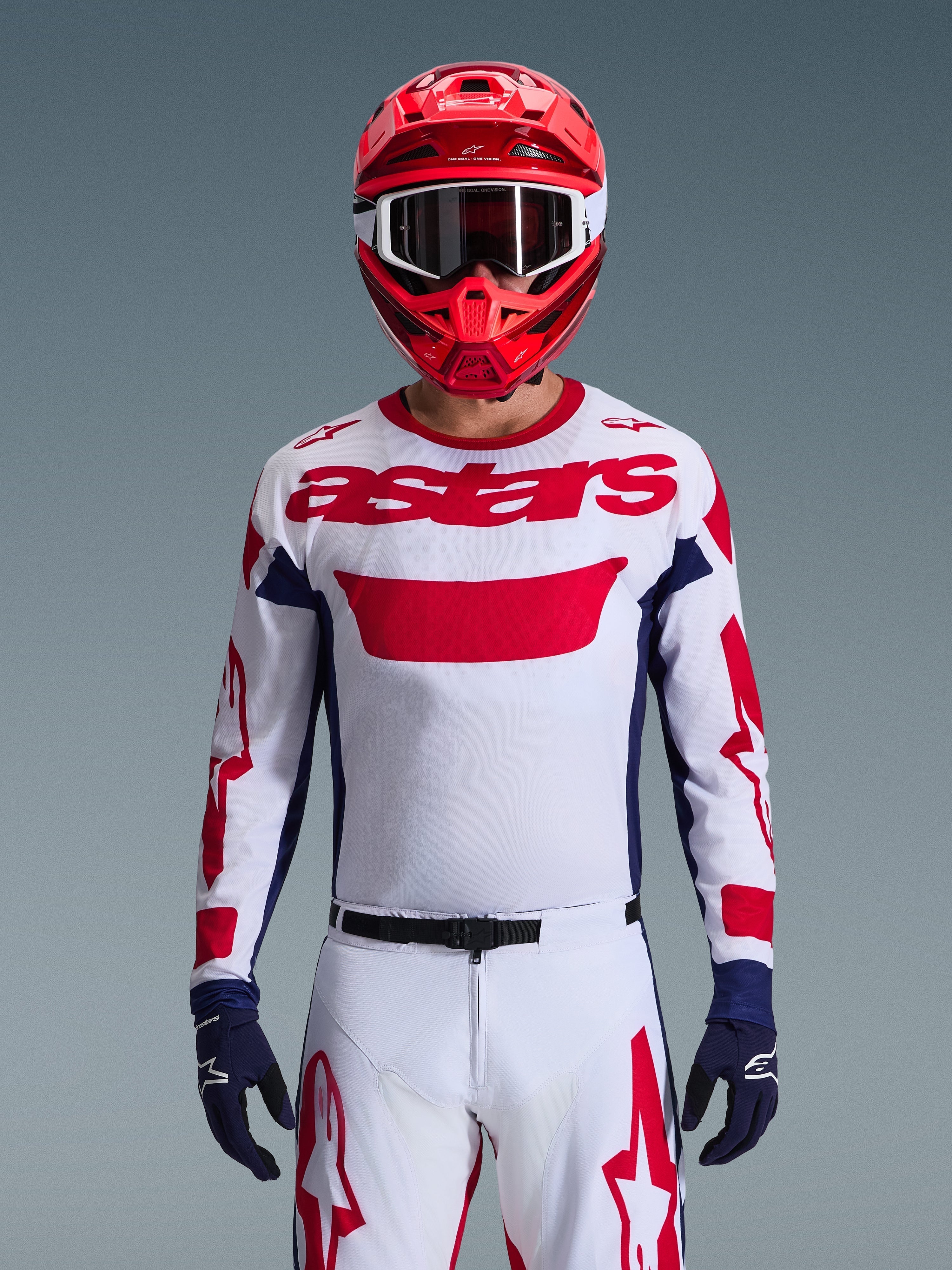 Zawodnik motocrossowy w koszulce Alpinestars 2026 Racer Riway w kolorystyce biało-niebiesko-czerwonej, duże czerwone logo „astars” na piersi, granatowe panele boczne, dopasowane biało-czerwone spodnie, czerwony kask z białymi goglami i granatowe rękawice, kompletny zestaw wyścigowy off-road