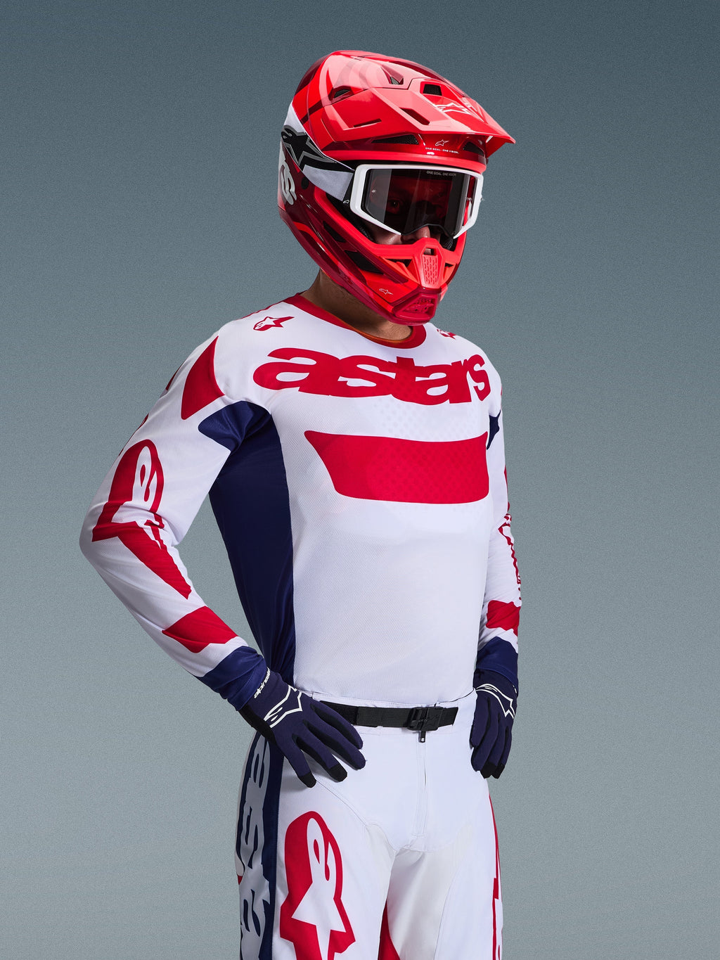 Zawodnik motocrossowy w koszulce Alpinestars 2026 Racer Riway w kolorze biało-niebiesko-czerwonym, z wyraźnym czerwonym brandingiem „astars” na klatce piersiowej i niebieskimi panelami bocznymi, w połączeniu z dopasowanym czerwonym kaskiem i białymi spodniami wyścigowymi, tworzącymi kompletny wyczynowy wygląd off-road.