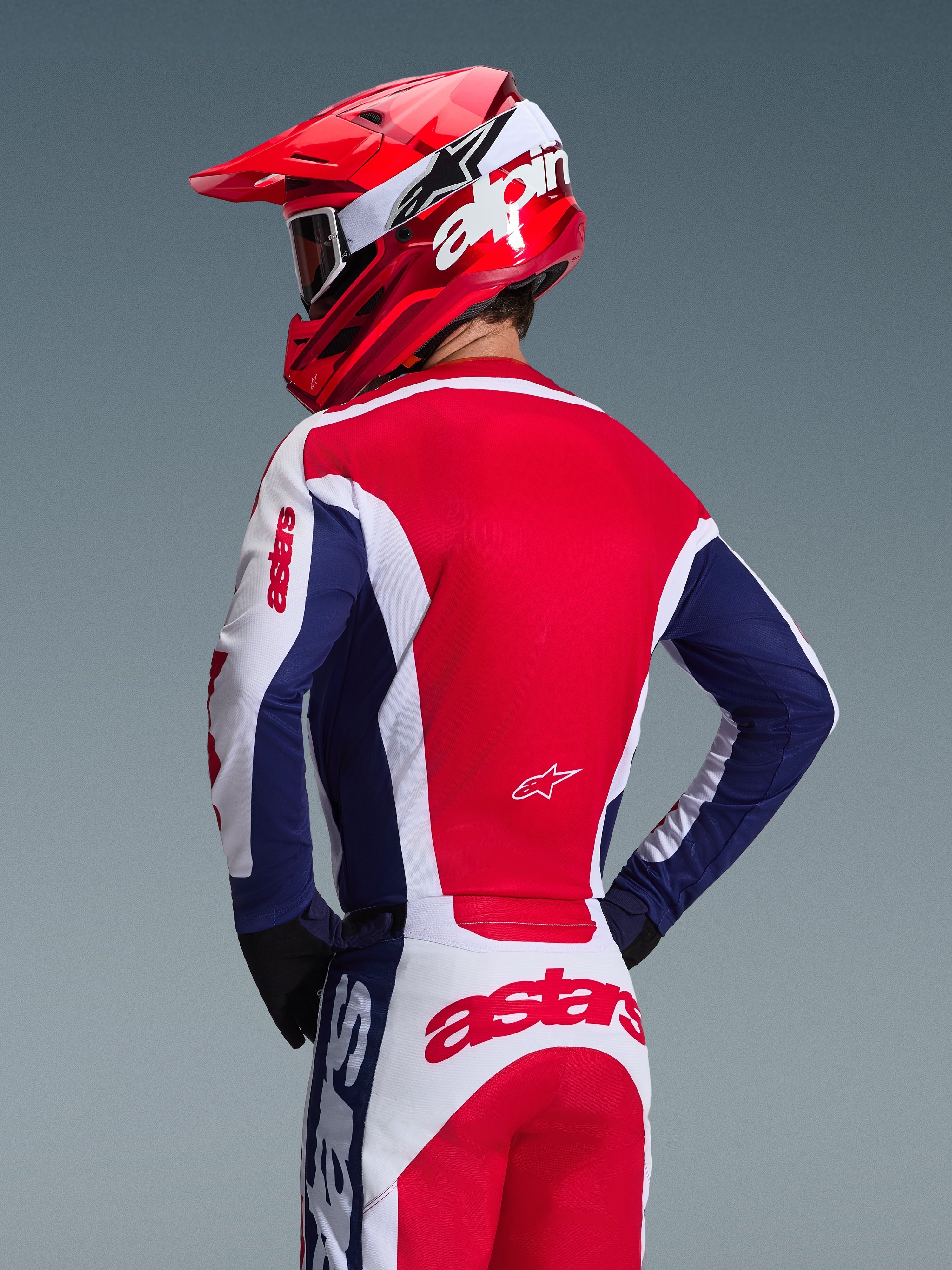 Motocyklista motocrossowy widziany od tyłu w koszulce Alpinestars 2026 Racer Riway, biało-niebiesko-czerwona kolorystyka, pasujące spodnie off-road z wyrazistym brandingiem astars, czerwony kask z białym logo Alpinestars, czarne rękawice, skoordynowany strój motocrossowy