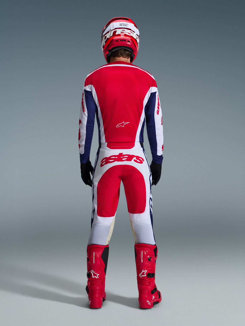 Motocyklista motocrossowy w pełnym stroju wyścigowym widoczny od tyłu, koszulka Alpinestars 2026 Racer Riway w barwach biało-niebiesko-czerwonych, pasujące biało-czerwone spodnie z wyraźnym brandingiem „astars”, czerwone buty oraz czerwono-biały kask, skoordynowany zestaw do jazdy off-road