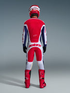 Motocyklista motocrossowy w pełnym stroju wyścigowym widoczny od tyłu, koszulka Alpinestars 2026 Racer Riway w barwach biało-niebiesko-czerwonych, pasujące biało-czerwone spodnie z wyraźnym brandingiem „astars”, czerwone buty oraz czerwono-biały kask, skoordynowany zestaw do jazdy off-road