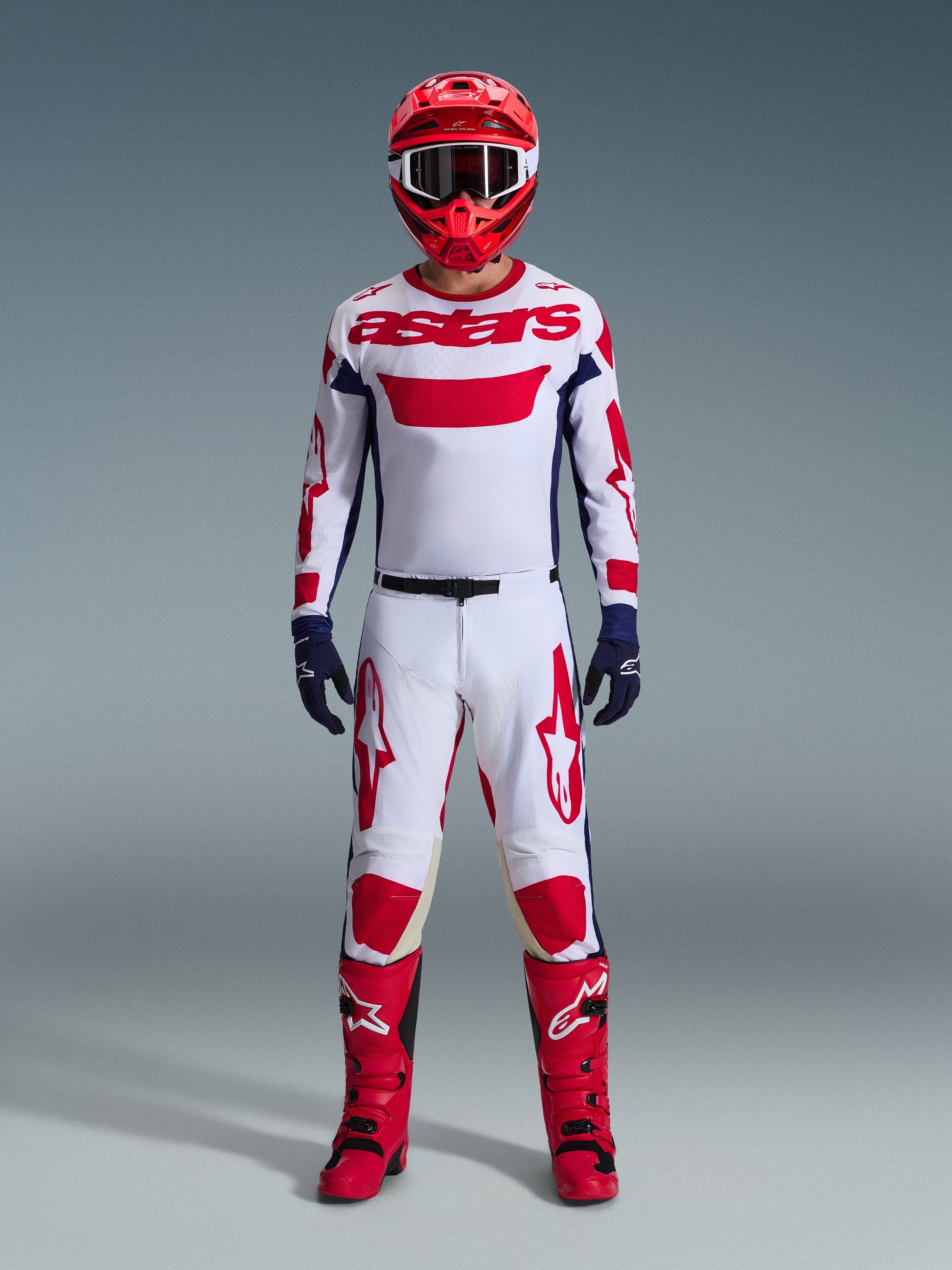 Zawodnik motocrossowy w pełnym stroju wyścigowym, koszulka Alpinestars 2026 Racer Riway w kolorze biało-niebiesko-czerwonym, pasujące biało-czerwone spodnie, czerwony kask, czerwone buty i granatowe rękawice, skoordynowany zestaw do wyścigów off-road