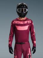 Kierowca motocrossowy w zestawie Alpinestars 2026 Racer Riway, bordowy i różowy, koszulka z wyrazistym różowym brandingiem na piersi, pasujące spodnie z czarnymi i różowymi akcentami, czarny kask full-face z goglami i czarne rękawice, spójny strój do wyścigów terenowych.