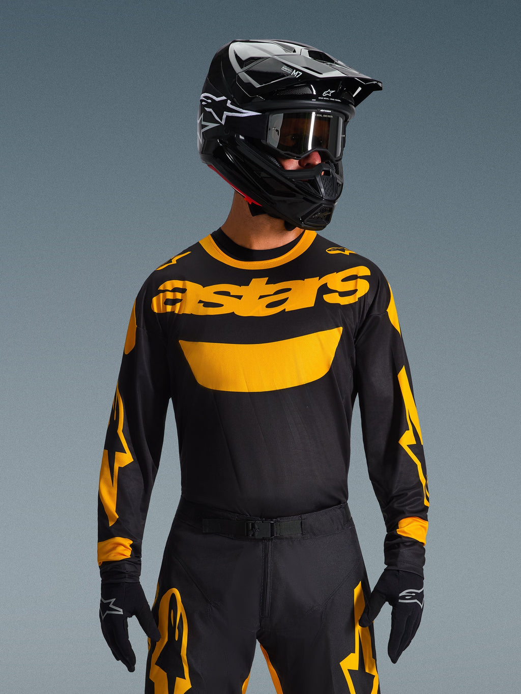 Motocrossowiec ubrany w koszulkę Alpinestars 2026 Racer Riway w kolorze żółto-czarnym, z wyraźnym żółtym logo „astars” na klatce piersiowej, w zestawie z dopasowanymi czarno-żółtymi spodniami, czarnym kaskiem z goglami oraz czarnymi rękawicami, profesjonalny zestaw odzieży off-road.