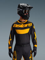 Motocrossowiec ubrany w koszulkę Alpinestars 2026 Racer Riway w kolorze żółto-czarnym, z wyraźnym żółtym logo „astars” na klatce piersiowej, w zestawie z dopasowanymi czarno-żółtymi spodniami, czarnym kaskiem z goglami oraz czarnymi rękawicami, profesjonalny zestaw odzieży off-road.