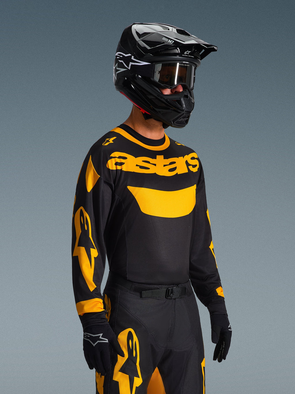 Kierowca motocrossowy w koszulce Alpinestars 2026 Racer Riway w barwach żółto-czarnych, czarna koszulka z wyraźnym żółtym brandingiem „astars” i grafikami na rękawach, pasujące czarno-żółte spodnie, czarny kask z goglami, czarne rękawice, profesjonalny zestaw wyścigowy off-road