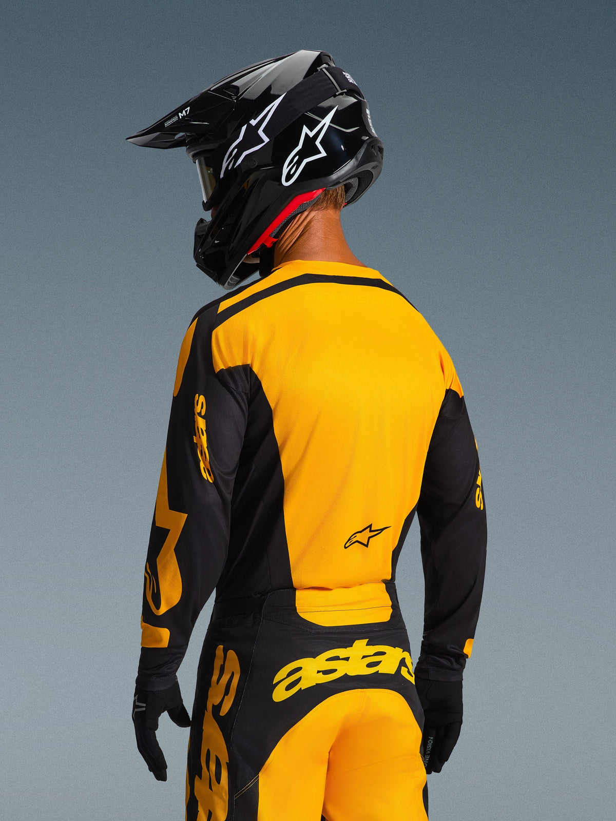 Widok z tyłu motocyklisty motocrossowego w koszulce Alpinestars 2026 Racer Riway w kolorze żółto-czarnym, z wyrazistym żółtym panelem tylnym i czarnymi rękawami z żółtym brandingiem, w zestawieniu z dopasowanymi spodniami motocrossowymi i czarnym kaskiem, tworzącymi kompletny zestaw wyścigowy off-road.