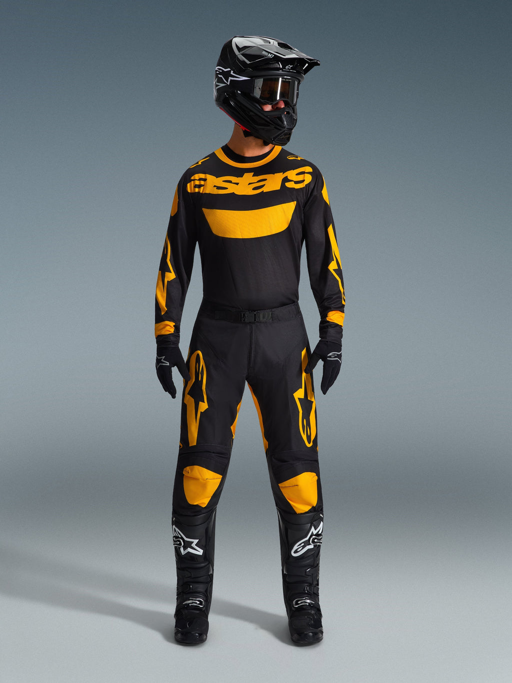Zawodnik motocrossowy w koszulce Racer Riway 2026 w kolorze żółto-czarnym, dopasowane spodnie, czarny kask oraz czarne buty z białym logo Alpinestars, pełny zestaw odzieży do wyścigów off-road.