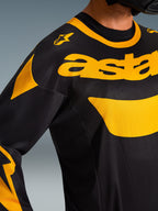 Zbliżenie na koszulkę Alpinestars 2026 Racer Riway, czarno-żółtą, z wyraźnym żółtym oznakowaniem na klatce piersiowej, techniczna konstrukcja z siateczki zapewniająca wentylację, profesjonalna odzież motocrossowa
