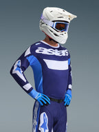 Motocrossista w koszulce Alpinestars 2026 Racer Riway w kolorze UCLA Blue White, z brandingiem „astars” na klatce piersiowej, w zestawie z niebieskimi spodniami motocrossowymi, białym kaskiem z goglami i niebieskimi rękawicami, wyczynowy sprzęt do jazdy terenowej