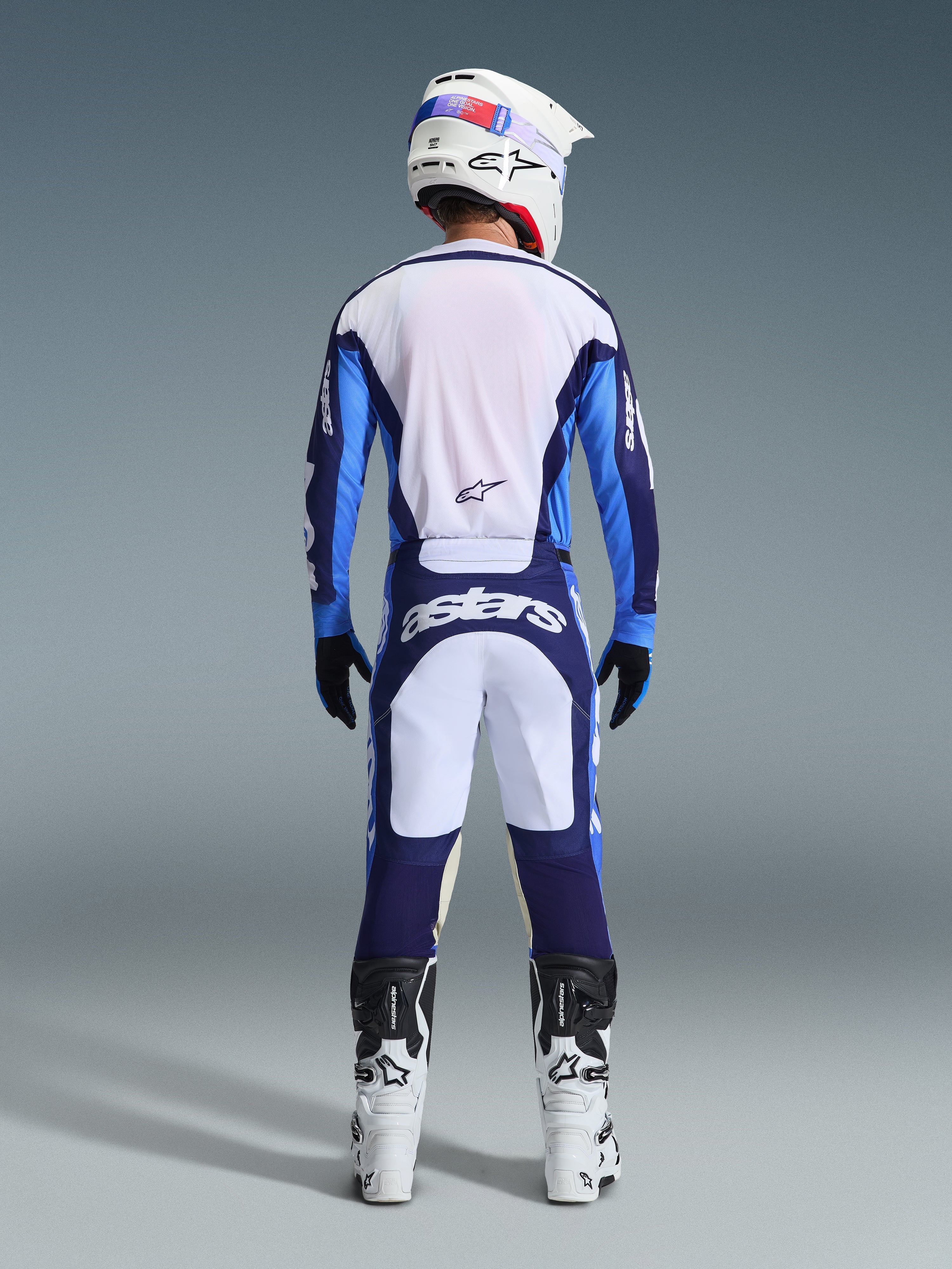 Rear view of a motocross rider wearing the Alpinestars 2026 Racer Riway Jersey and Pants in Ucla Blue and White, coordinated with a white helmet, black gloves, and white motocross boots, high-performance gear for off-road racing