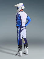 Motocyklista motocrossowy w koszulce 2026 Racer Riway w kolorze niebiesko-białym (Ucla Blue White), w komplecie z dopasowanymi spodniami, białym kaskiem i białymi butami, widok z tyłu pod kątem trzech czwartych, kompletny zestaw wyścigowy off-road.