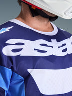 Zbliżenie na koszulkę Alpinestars 2026 Racer Riway w kolorach niebieskim Ucla i białym, z dużym białym logo „astars” na piersi, białym kołnierzykiem i oddychającą teksturą materiału typu mesh, noszoną z białym kaskiem motocrossowym.