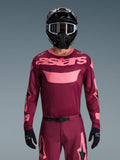 Motocyklista motocrossowy w koszulce Alpinestars 2026 Racer Riway w kolorze Dark Red Coral, z bordowym korpusem i koralowo-różowym logotypem „astars” na piersi, w zestawie z pasującymi spodniami motocrossowymi, czarnym kaskiem i czarnymi rękawicami, profesjonalny zestaw odzieży do jazdy off-road.