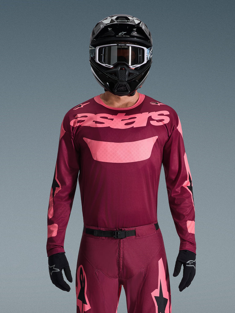 Motocyklista motocrossowy w koszulce Alpinestars 2026 Racer Riway w kolorze Dark Red Coral, z bordowym korpusem i koralowo-różowym logotypem „astars” na piersi, w zestawie z pasującymi spodniami motocrossowymi, czarnym kaskiem i czarnymi rękawicami, profesjonalny zestaw odzieży do jazdy off-road.