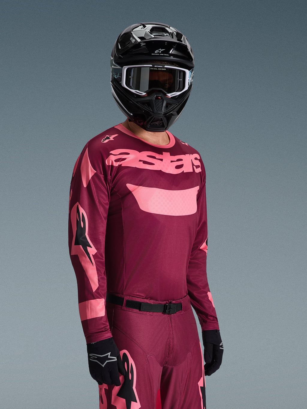 Motocyklista motocrossowy ubrany w koszulkę Alpinestars 2026 Racer Riway w kolorze Dark Red Coral i dopasowane spodnie, koralowo-różowy branding „astars” na piersi, czarny kask i czarne rękawice, skoordynowany zestaw odzieży wyścigowej off-road