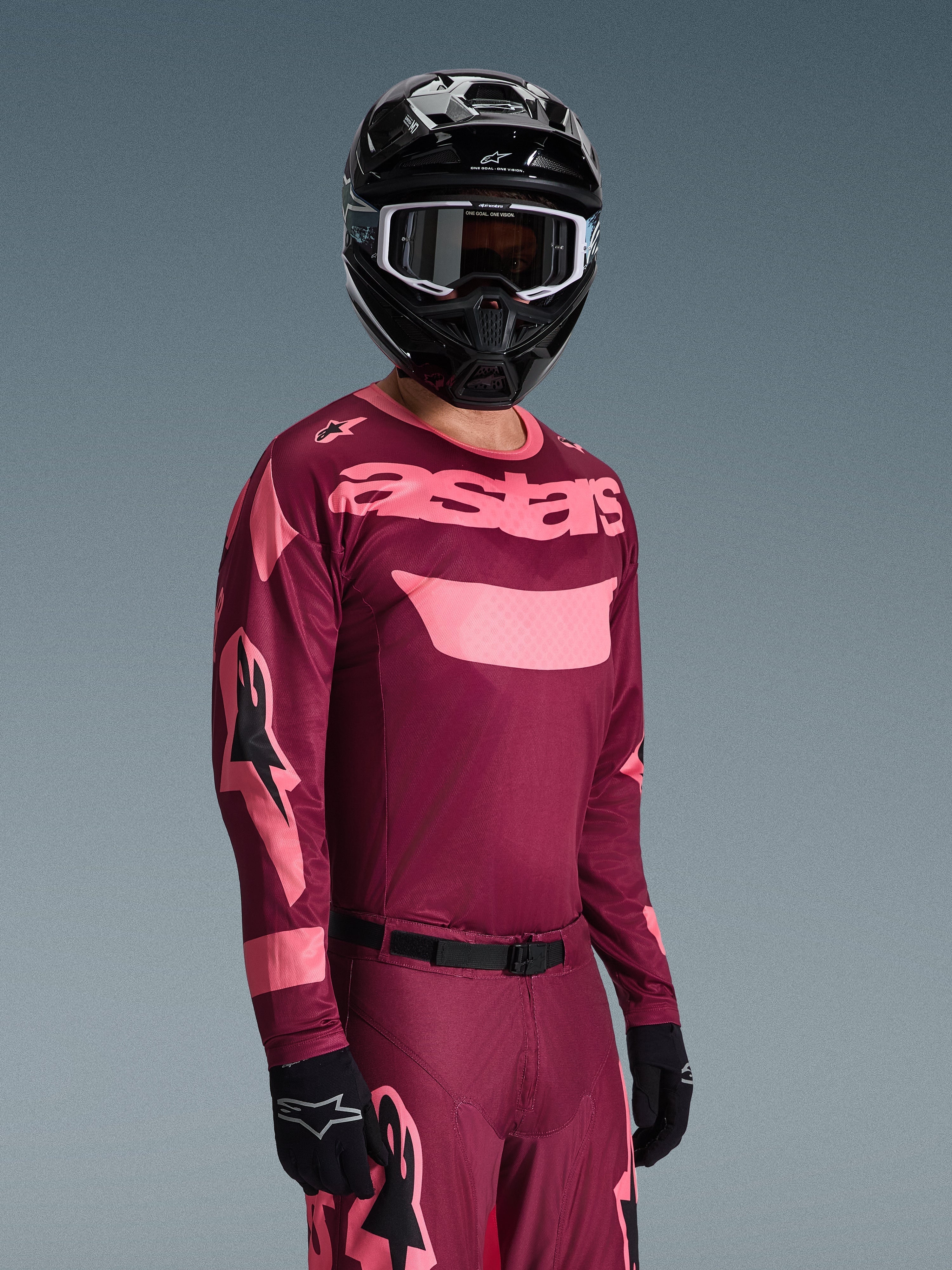 Motocyklista motocrossowy ubrany w koszulkę Alpinestars 2026 Racer Riway w kolorze Dark Red Coral i dopasowane spodnie, koralowo-różowy branding „astars” na piersi, czarny kask i czarne rękawice, skoordynowany zestaw odzieży wyścigowej off-road